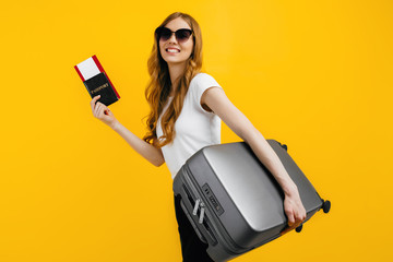 Smiling female passenger in sunglasses, standing with a suitcase and passport with tickets, on a yellow background. Travel concept