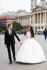 Naklejka premium Bride and groom dancing in the old town street. Wedding couple walks in Budapest near Parliament House. Caucasian happy romantic young couple celebrating their marriage. Wedding and love concept.