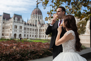 Bride and groom have fun and eating ice cream. Wedding couple sit in Budapest near Parliament House. Caucasian happy romantic young couple celebrating their marriage. Wedding and love concept.