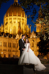 Bride and groom hugging in the old town street. Wedding couple walks in Budapest near Parliament House in night. Happy romantic young couple celebrating their marriage. Wedding and love concept.