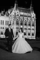 Obraz premium Bride and groom hugging in the old town street. Wedding couple walks in Budapest near Parliament House in night. Happy romantic young couple celebrating their marriage. Wedding and love concept.