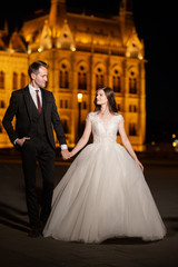 Naklejka premium Bride and groom hugging in the old town street. Wedding couple walks in Budapest near Parliament House in night. Happy romantic young couple celebrating their marriage. Wedding and love concept.