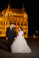 Obraz premium Bride and groom hugging in the old town street. Wedding couple walks in Budapest near Parliament House in night. Happy romantic young couple celebrating their marriage. Wedding and love concept.