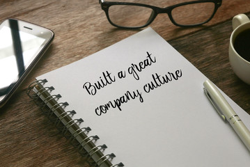 Selective focus of mobile phone,glasses,a cup of coffee,pen and notebook written with ' Built a great company culture ' on wooden background.