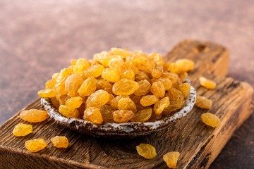 Organic dried golden raisins in ceramic plate on wooden board. Brown background, close up.