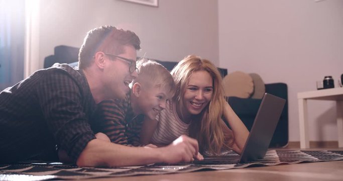 Happy family young parents couple and cute kid son using laptop looking at computer screen enjoying watching funny social media video doing online shopping relaxing on carpet at home together