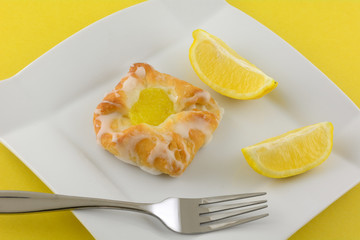 Mini lemon danish breakfast pastry with fresh lemon wedges on white plate with fork on yellow background