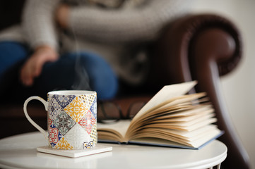 Staying at home: woman sitting on a classic vintage leather sofa relaxing with a cup of herbal tea (infusion) and an open book on a small table in front of her.