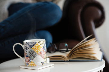 Staying at home: woman sitting on a classic vintage leather sofa relaxing with a cup of herbal tea (infusion) and an open book on a small table in front of her.