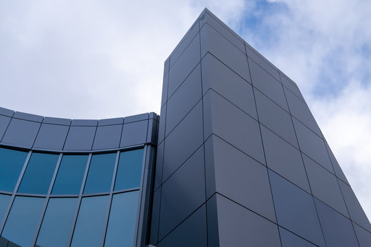 The corner of a grey coloured building made of metal composite panels and green reflective windows.   Part of the building has a high tower and a lower curved section made of green windows. 