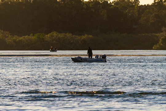 A silhouetted fisherman in a small fishing boat casts his fishing line near a sandbar on the Noosa River in the late afternoon.