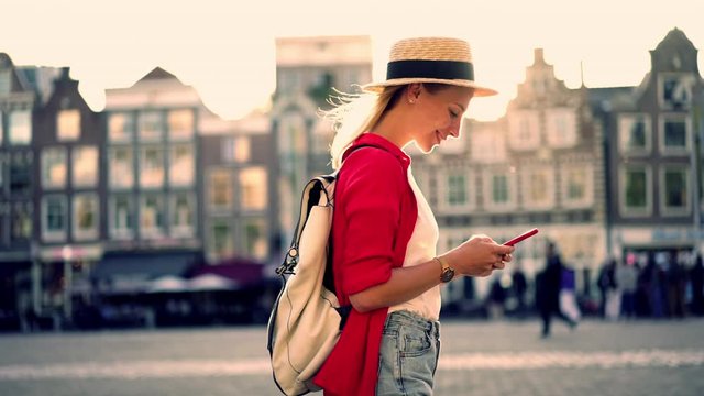 Happy female tourist walking in Amsterdam city while sending media message sharing photos of sightseeing.  carefree hipster girl chatting in mobile phone while enjoying vacations at sunset,slow motion