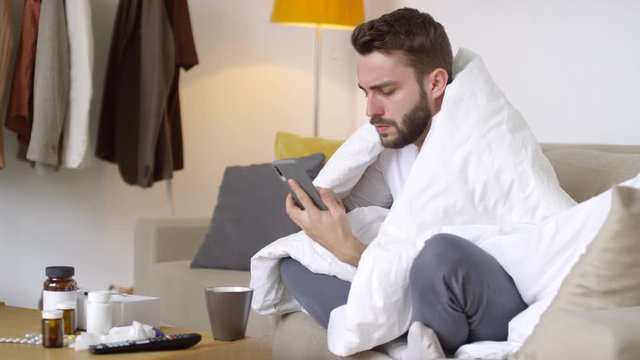 Medium shot of sick young man wrapped in blanket taking mobile phone and calling his doctor to ask something about his prescription drugs