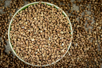 Buckwheat is poured in a round bowl (plate), close-up top view, around defocused buckwheat scattered around the table