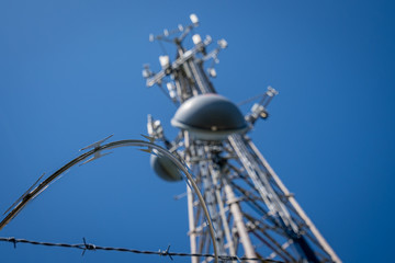 Cellphone tower with communication dishes and transmission antennas, surrounded by barbed and razor wire. against a clear blue sky.