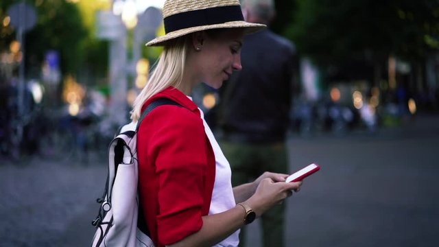 Happy hipster girl with stylish backpack enjoying time for walking across showplaces during installing application for tracking gps on cellular phone, positive female tourist using smartphone app 
