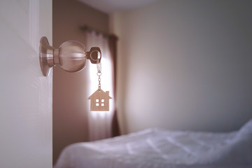 Half opened door to bedroom and house key with house wood shaped keyring in lock door. Sunlight passing through window hits to white bed in  bedroom. Opening door Welcome, Entrance to bedroom concept