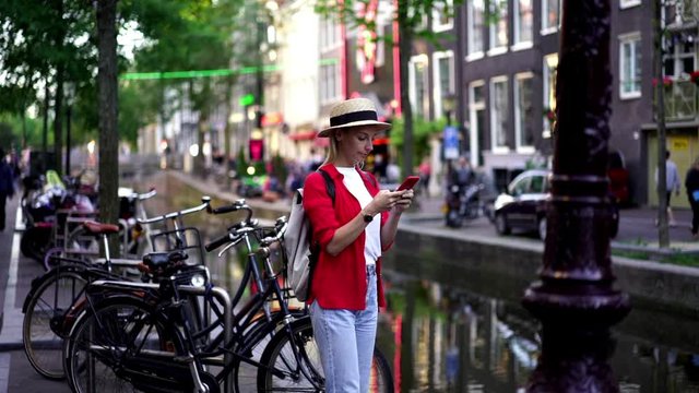Slow motion of positive hipster girl standing in Amsterdam city near canal and modern bicycles using 4g internet in roaming on mobile phone. Millennial female tourist messaging on smartphone outdoors
