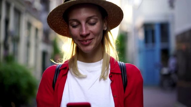 Slow motion of happy hipster girl in hat walking across sunny city streets and downloading media files from mobile application for making publication in social networks connected to wireless internet
