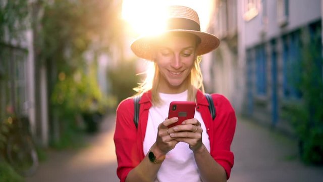Slow motion of positive smiling hipster girl in hat happy of received message with good news, Cheerful young woman using modern technology while browsing internet. Soft focus on smartphone in hands
