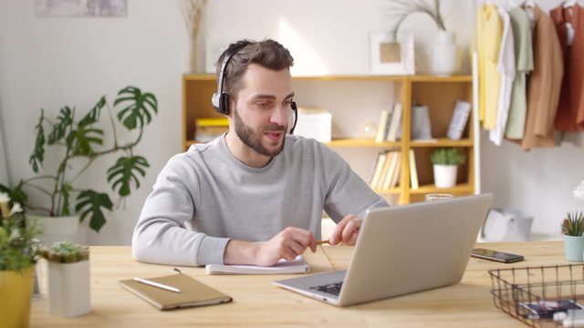 Medium shot of cheerful young man in wireless headset talking on video call on laptop while working remotely from home, then ending call and smiling for camera