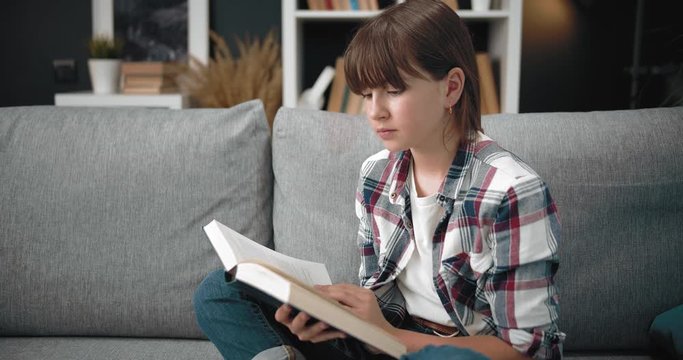 Pretty young girl in casual clothing sitting on grey couch and reading interesting book. Female teenager with dark hair spending free time at home for education.