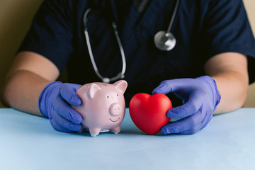 A medical professional with gloved hands holding a pink piggy bank and a red heart
