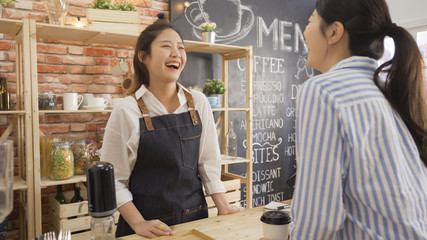 cheerful female waiter taking order from young female costumer standing at cafe counter. two laughing women friends meeting in coffee shop while one working as waitress. happy regular client indoors