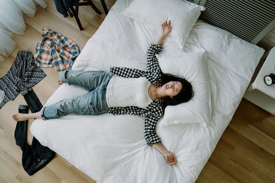 top view of taiwanese woman lying in a sprawl on bed with her clothes scattered on floor. asian young girl hanging out with friend until dawn goes to bed straight without taking bath.