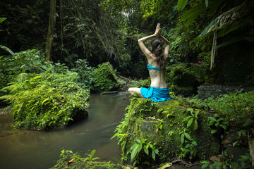 Young Caucasian woman sitting on the rock, practicing yoga and pranayama. Raising arms with namaste...