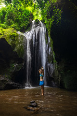 Young Caucasian woman standing near the waterfall, meditating, practicing yoga and pranayama. Hands raising up with namaste mudra. Krisik waterfall in Bangli, Bali, Indonesia.