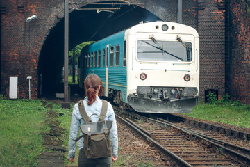 Blue passenger train rides from brown tunnel. Random passer girl traveler with backpack is walking...
