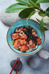 Poke bowl with salmon fillet on a light-blue stone surface with pebble stones and green ficus leaves, vertical shot