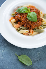 Penne pasta with bolognese sauce and fresh green basil in a white plate, close-up on a blue stone surface, vertical shot
