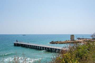 Fototapeta na wymiar People walking on a pier with statue of Poseidon, bridge near the beach, blue sea and sky