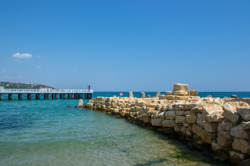 Fototapeta na wymiar People walking on a pier with statue of Poseidon, bridge near the beach, blue sea and sky
