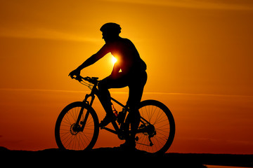 Fototapeta premium Silhouette of a biker on sky background on sunset. High hill top and wide river on orange sunset.
