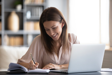 Smiling woman writing notes in notebook daily planner, planning week, working or studying at home, using laptop, beautiful female wearing wireless earphones listening to music or lecture