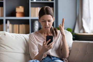 Confused angry woman having problem with phone, sitting on couch at home, unhappy young female...