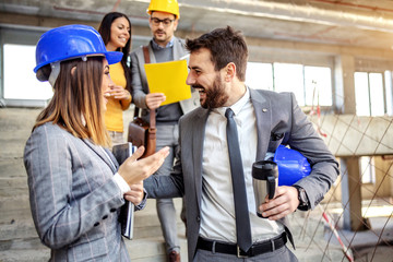 Four dedicated architects standing in building in construction process and talking about important project. Man and woman in foreground chatting while other ones talking about ides.