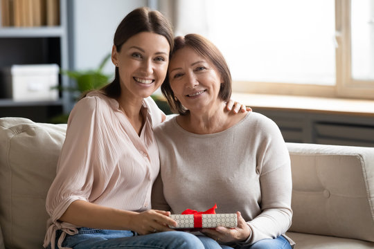 Portrait Daughter Hugging And Congratulating Older Mother With Birthday, Mothers Day Or 8 March, Happy Young Woman And Mature Mom Holding Gift Box, Looking At Camera, Sitting On Couch At Home