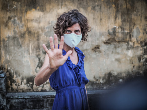 woman standing in front of a vintage wall wearing a face mask protecting from corona virus covid-19 holding up hand saying stop 