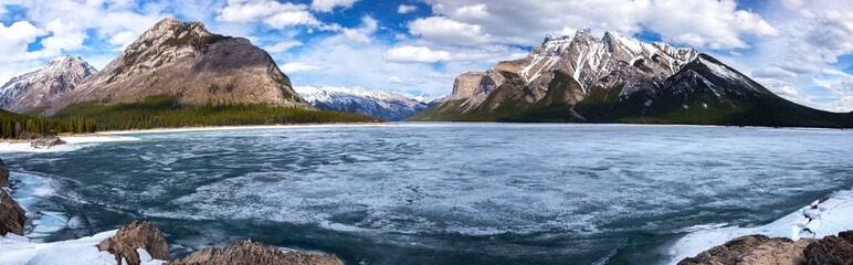 Fototapeta na wymiar Panoramic Landscape View of Frozen Lake Minnewanka Surface in Springtime, Alberta Rocky Mountains. Vehicle Access to Canadian National Parks has been closed because of COVID-19 Virus Pandemic