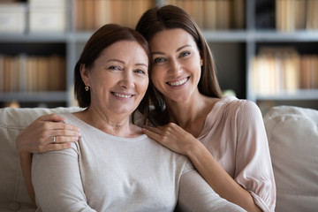 Head shot portrait smiling mature mother and daughter hugging, sitting on couch at home, looking at...