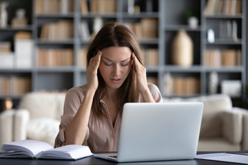 Naklejka premium Tired woman massaging temples, suffering from headache after computer work, sitting at desk with laptop at home, exhausted young female with closed eyes touching head, relieving pain, migraine