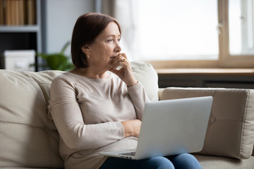 Thoughtful mature woman using laptop, sitting on couch at home alone, pensive older female thinking about finance or health problem, looking in distance, having doubts, feeling lonely