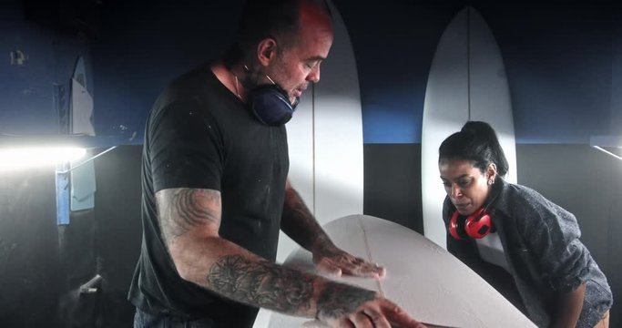 Man shaping surfboard and black woman analysing shape in workshop