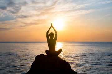 girl is sitting in lotus pose on the big rock in front of sunrise at the sea