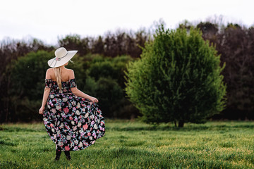 a beautiful girl in a dress and a hat walks alone in a green meadow. Young pretty woman blonde outdoors enjoy freedom and relaxation