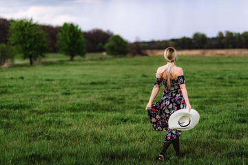a beautiful girl in a dress with a hat in her hand walks alone in a green meadow. Young pretty blond woman in nature runs forward and enjoys freedom and relaxation